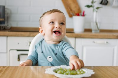Charming little baby boy eating first food green grape at the bright kitchen at home